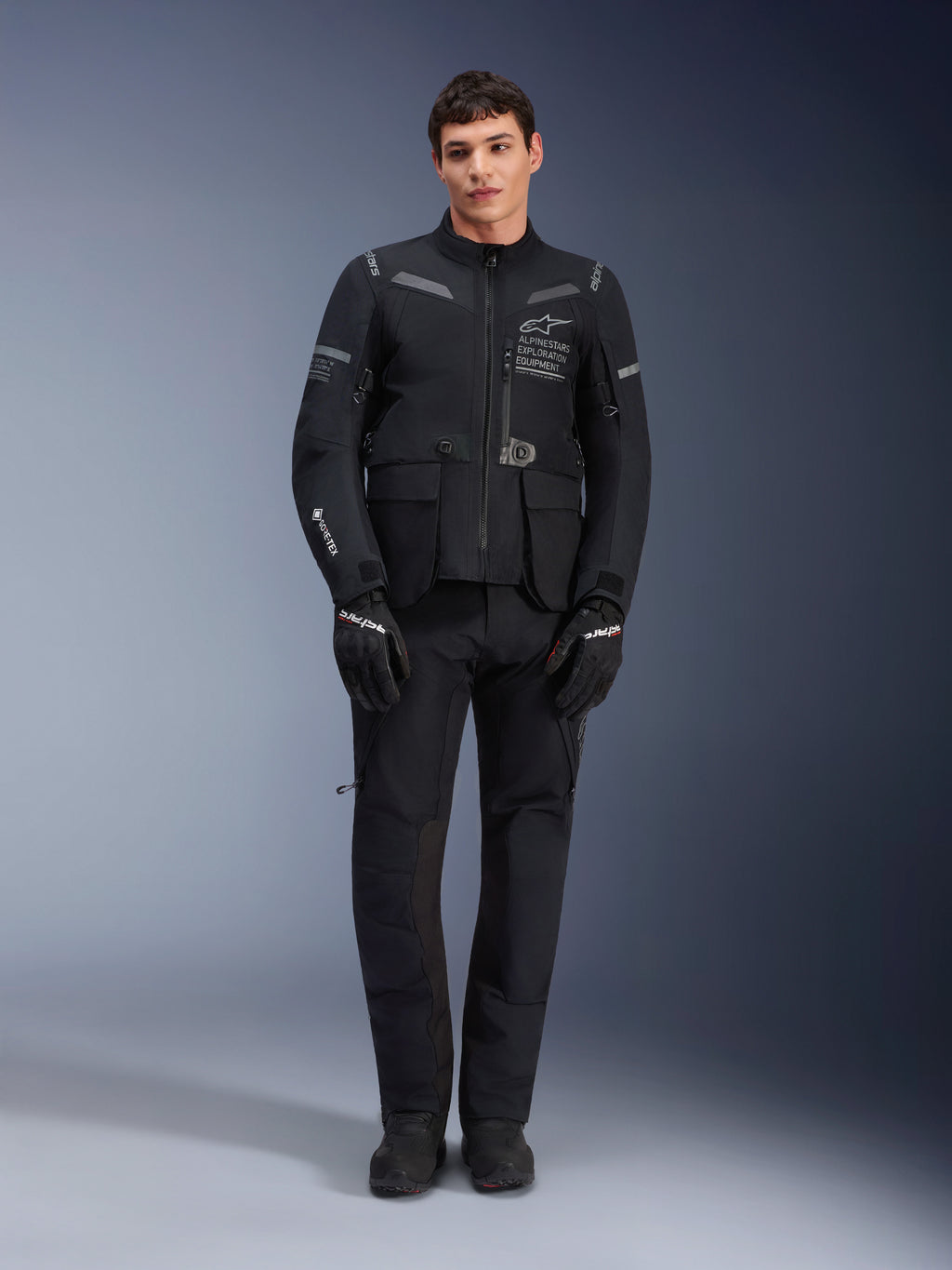 Alpinestars ST-7 2L GORE-TEX Jacket, adventure touring motorcycle jacket, Black and Dark Gray, worn with matching black riding pants and gloves, featuring multiple cargo pockets, reflective accents, and Alpinestars Exploration Equipment chest branding, functional design for road and off-road riding