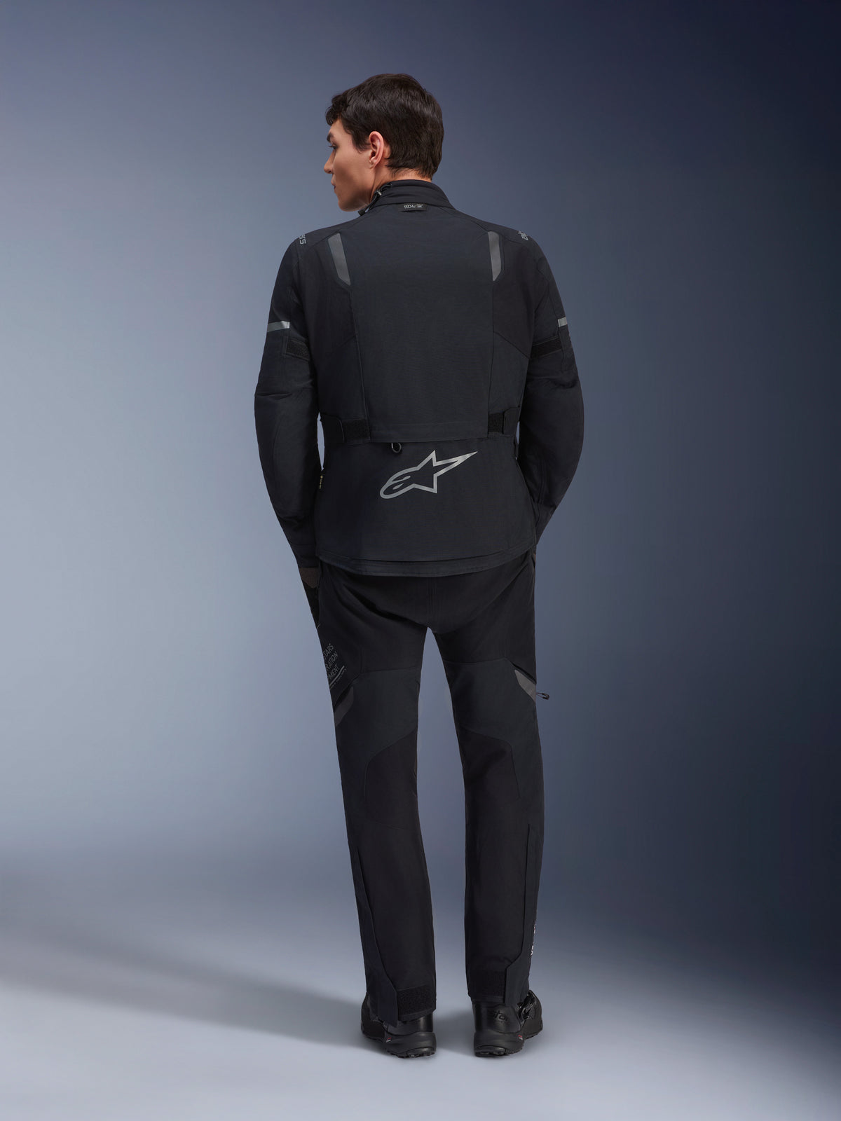 Alpinestars ST-7 2L GORE-TEX Jacke und passende ST-7 2L GORE-TEX Hose, schwarz-dunkelgraue Adventure-Motorradausrüstung, Rückansicht mit reflektierendem Alpinestars-Logo auf der Jacke und technischer Textilkonstruktion.