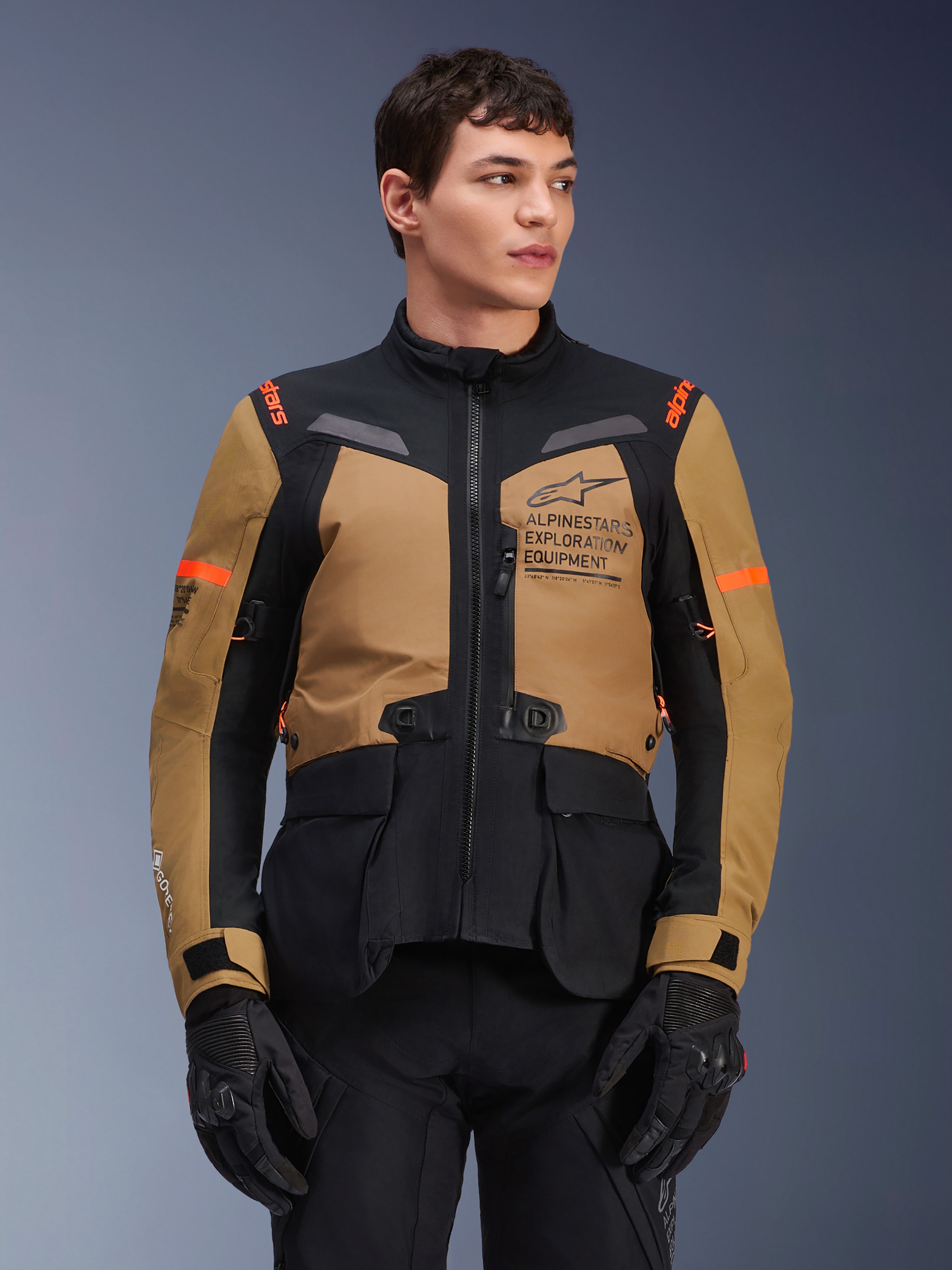 Alpinestars ST-7 2L GORE-TEX Jacke, Motorrad-Adventure-Touringjacke, Gobi-Braun und Schwarz mit orangen Akzenten, getragen von einem männlichen Model, mit Exploration Equipment Branding und GORE-TEX-Technologie, strapazierfähige wasserdichte Fahrausrüstung