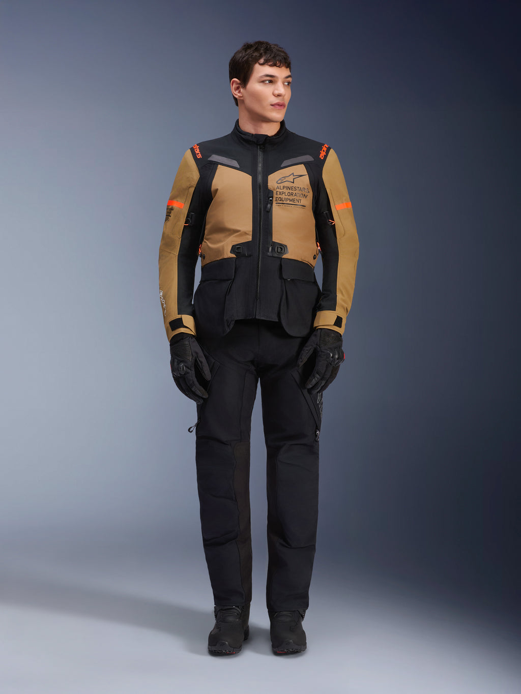 Männliches Model in Alpinestars ST-7 2L GORE-TEX Jacke, Gobi-Braun und Schwarz, Adventure-Motorradausrüstung mit passender schwarzer Hose und Handschuhen, mit Alpinestars Exploration Equipment Branding auf der Brust, funktionales wasserdichtes Design für Straßen- und Offroad-Touren