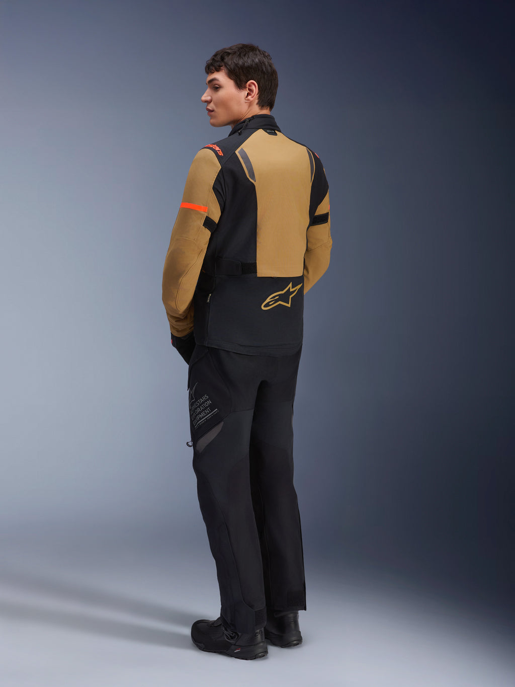 Alpinestars ST-7 2L GORE-TEX Jacke, Adventure-Motorradjacke, Gobi-Braun und Schwarz, Rückansicht eines Models in passender Fahrhose und Stiefeln, mit orangefarbenen Akzenten an den Ärmeln, wasserdichtem laminiertem Obermaterial und Alpinestars-Logo, entwickelt für Touren und Straßenfahrten