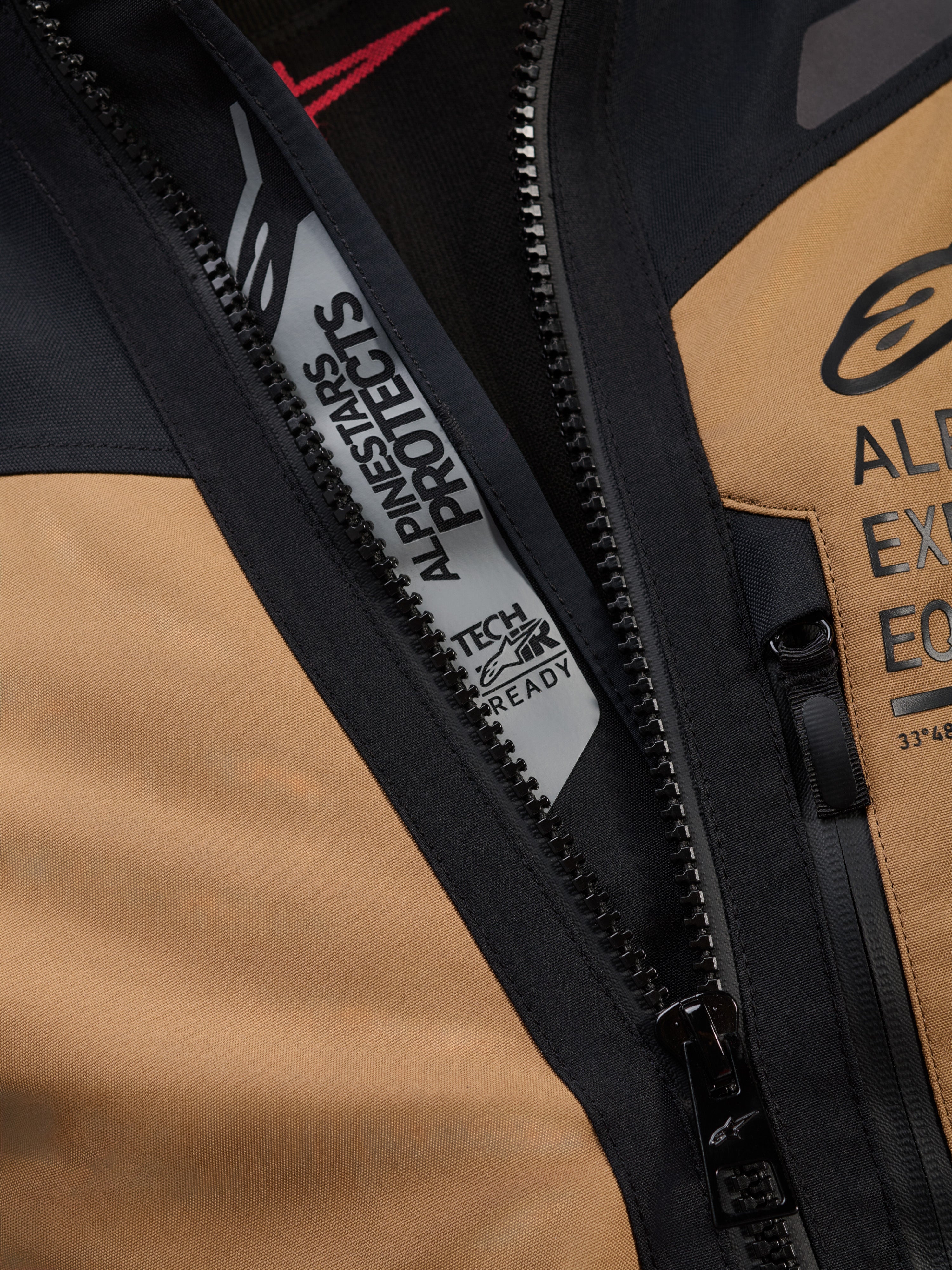 Alpinestars ST-7 2L GORE-TEX Jacke, Detailaufnahme in Gobi Braun und Schwarz, mit „ALPINESTARS PROTECTS“ und „TECH-AIR READY“ Markenzeichen an der Innenseite des Hauptreißverschlusses, robuste Textilkonstruktion und Logos auf der Brust