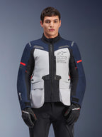 Alpinestars ST-7 2L GORE-TEX Jacke, Motorrad-Adventure-Touringjacke, Eisgrau und Dunkelblau mit roten Akzenten, wasserdicht und atmungsaktiv, getragen von einem Modell mit schwarzen Handschuhen, mit Alpinestars Exploration Equipment Branding auf der Brust