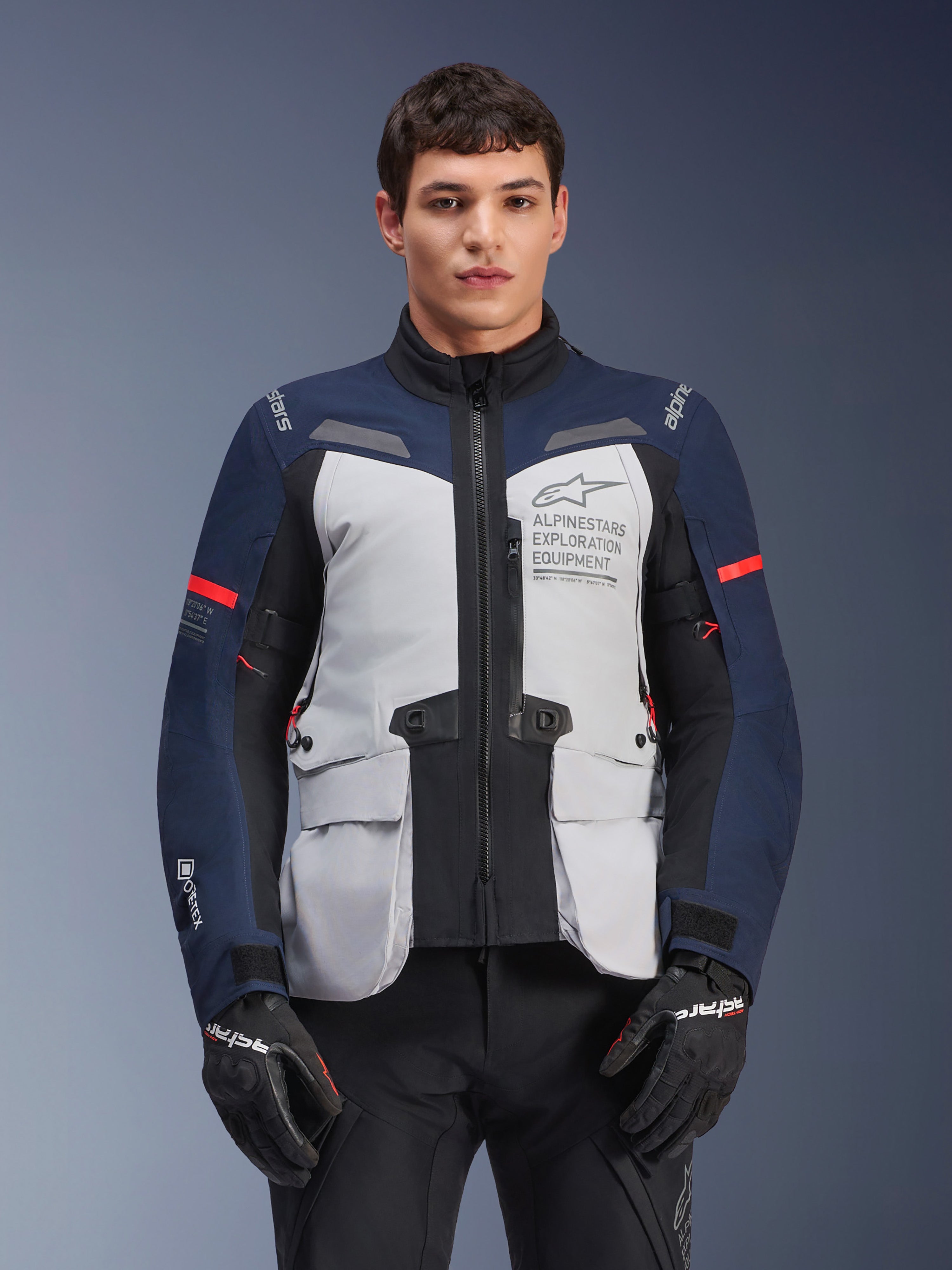 Alpinestars ST-7 2L GORE-TEX Jacke, Motorrad-Adventure-Touringjacke, Eisgrau und Dunkelblau mit roten Akzenten, wasserdicht und atmungsaktiv, getragen von einem Modell mit schwarzen Handschuhen, mit Alpinestars Exploration Equipment Branding auf der Brust