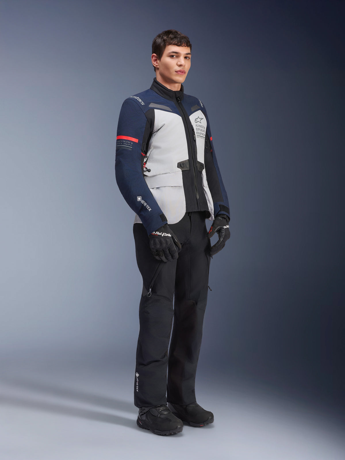 Alpinestars ST-7 2L GORE-TEX Jacke in Ice Gray, Dunkelblau und Schwarz mit roten Akzenten, getragen von einem männlichen Modell mit schwarzer Motorradhose und Handschuhen, professionelle Adventure-Touring-Motorradausrüstung