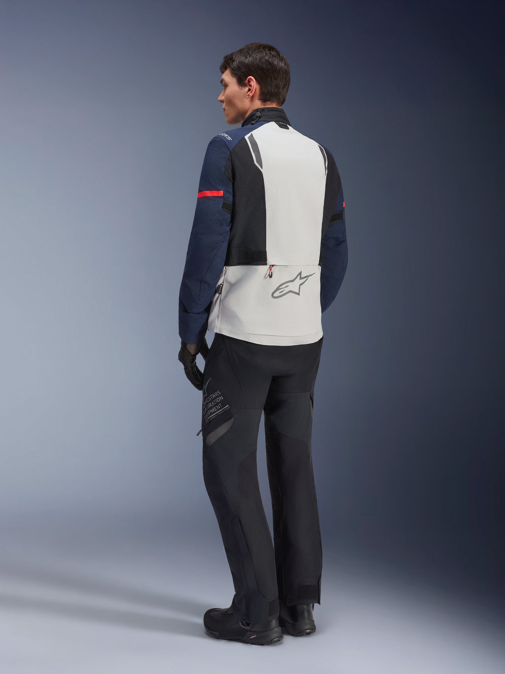 Model in der Alpinestars ST-7 2L GORE-TEX Jacke in Eisgrau, Dunkelblau und Schwarz, Rückansicht, hellgraues Rückenpanel mit Alpinestars-Logo, dunkelblaue Ärmel mit rotem Streifen, kombiniert mit schwarzer Adventure-Fahrhose und Stiefeln, Motorrad-Tourenbekleidung