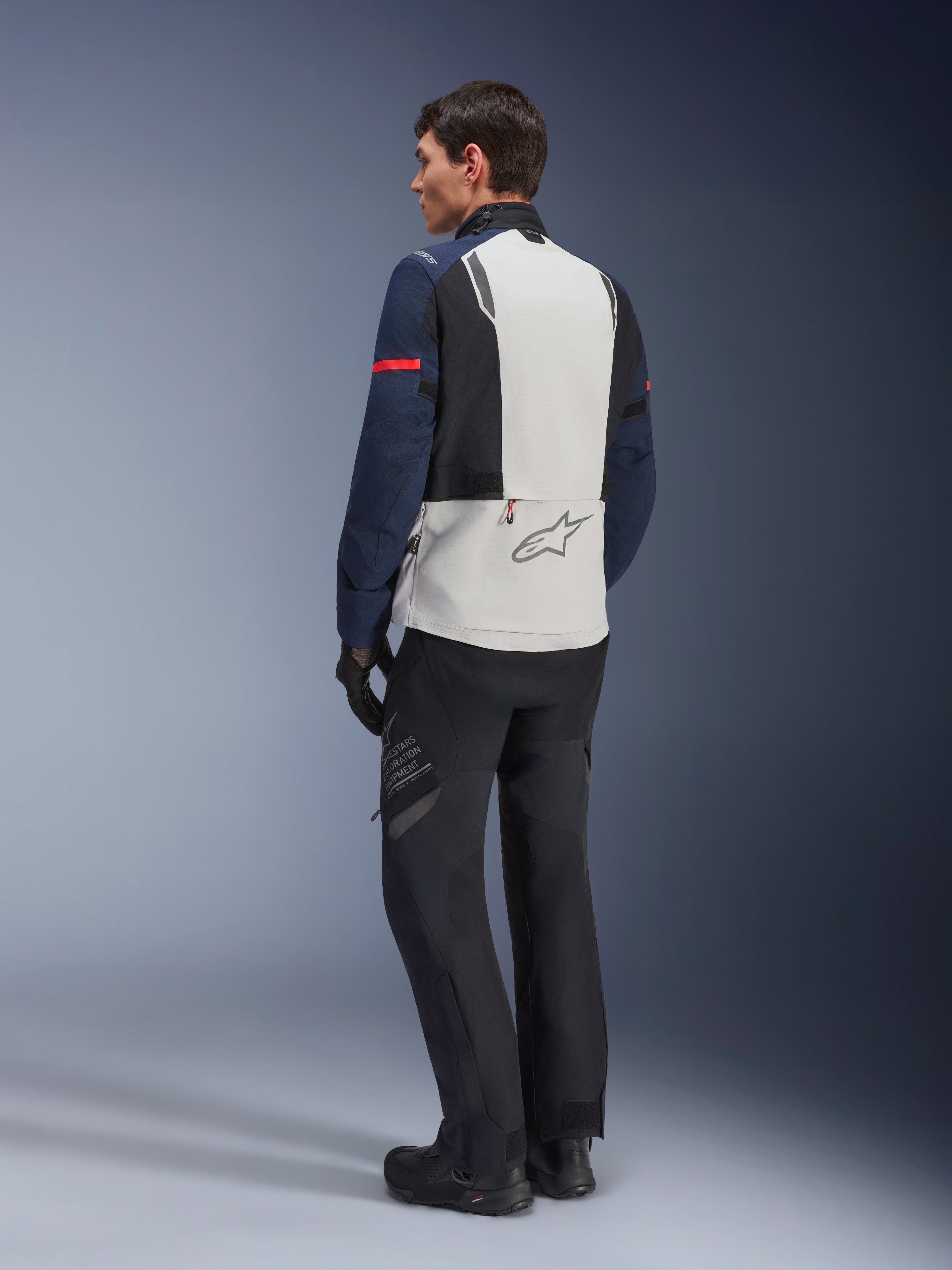 Model in der Alpinestars ST-7 2L GORE-TEX Jacke in Eisgrau, Dunkelblau und Schwarz, Rückansicht, hellgraues Rückenpanel mit Alpinestars-Logo, dunkelblaue Ärmel mit rotem Streifen, kombiniert mit schwarzer Adventure-Fahrhose und Stiefeln, Motorrad-Tourenbekleidung