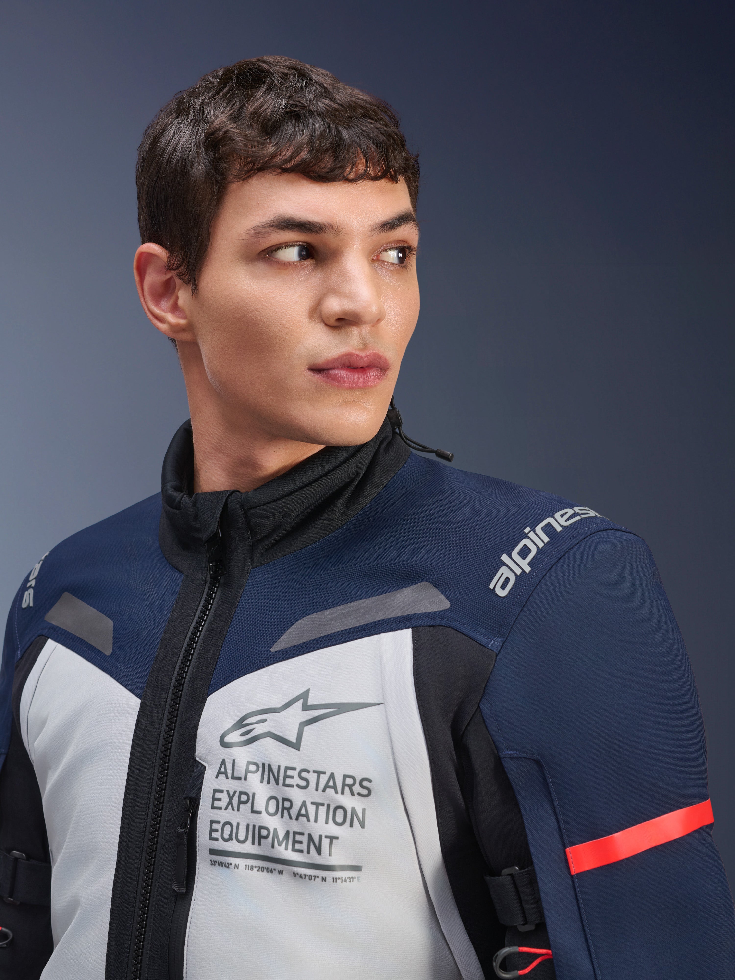 Alpinestars ST-7 2L GORE-TEX Jacke, Eisgrau, Dunkelblau und Schwarz, von einem männlichen Model getragen, mit Alpinestars Exploration Equipment-Branding auf der Brust, hohem Kragen und rotem Akzent am Ärmel, strapazierfähiges Textildesign für Adventure-Touren
