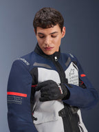 Alpinestars ST-7 2L GORE-TEX Jacke, Ice Gray, Dunkelblau und Schwarz mit orangefarbenen Akzenten, getragen von einem Mann, der ein Ärmeldetail anpasst, mit „Alpinestars Exploration Equipment“-Branding und geografischen Koordinaten, professionelle Adventure-Motorradbekleidung