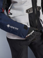 Alpinestars ST-7 2L GORE-TEX Jacke, Nahaufnahme eines dunkelblauen Ärmels mit weißem GORE-TEX Logo, eisgrauer Torso mit schwarzen Details und roten Kordelakzenten, kombiniert mit schwarzen Alpinestars Handschuhen und Hose, Adventure-Touring-Motorradausrüstung