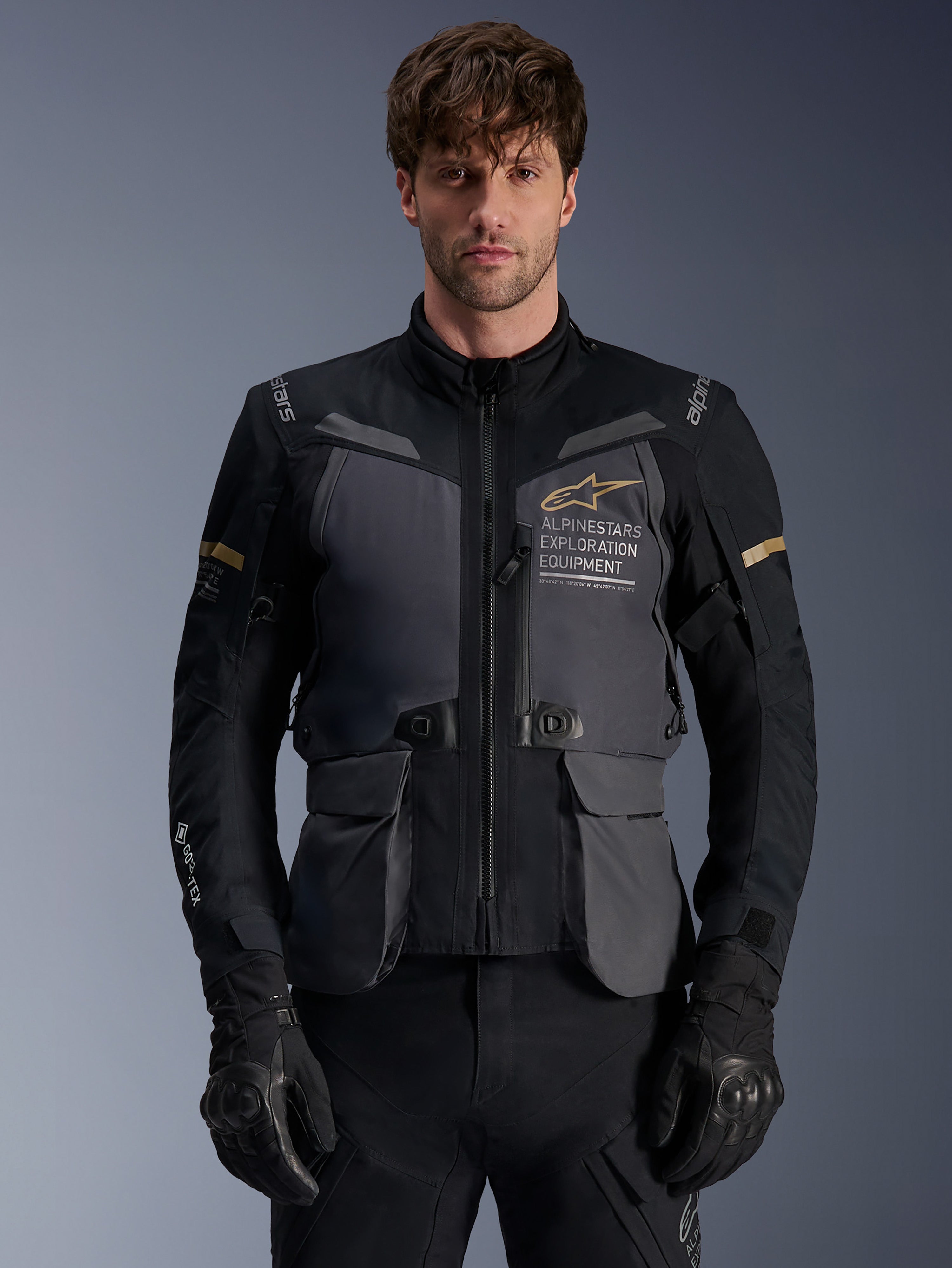 Alpinestars ST-7 2L GORE-TEX Jacke, Herren Adventure-Touring-Motorradjacke, Dunkelgrau und Schwarz mit khakifarbenen Akzenten, mit „Alpinestars Exploration Equipment“-Branding, wasserdichten Cargotaschen und hohem Kragen, getragen von einem Model mit schwarzen Handschuhen