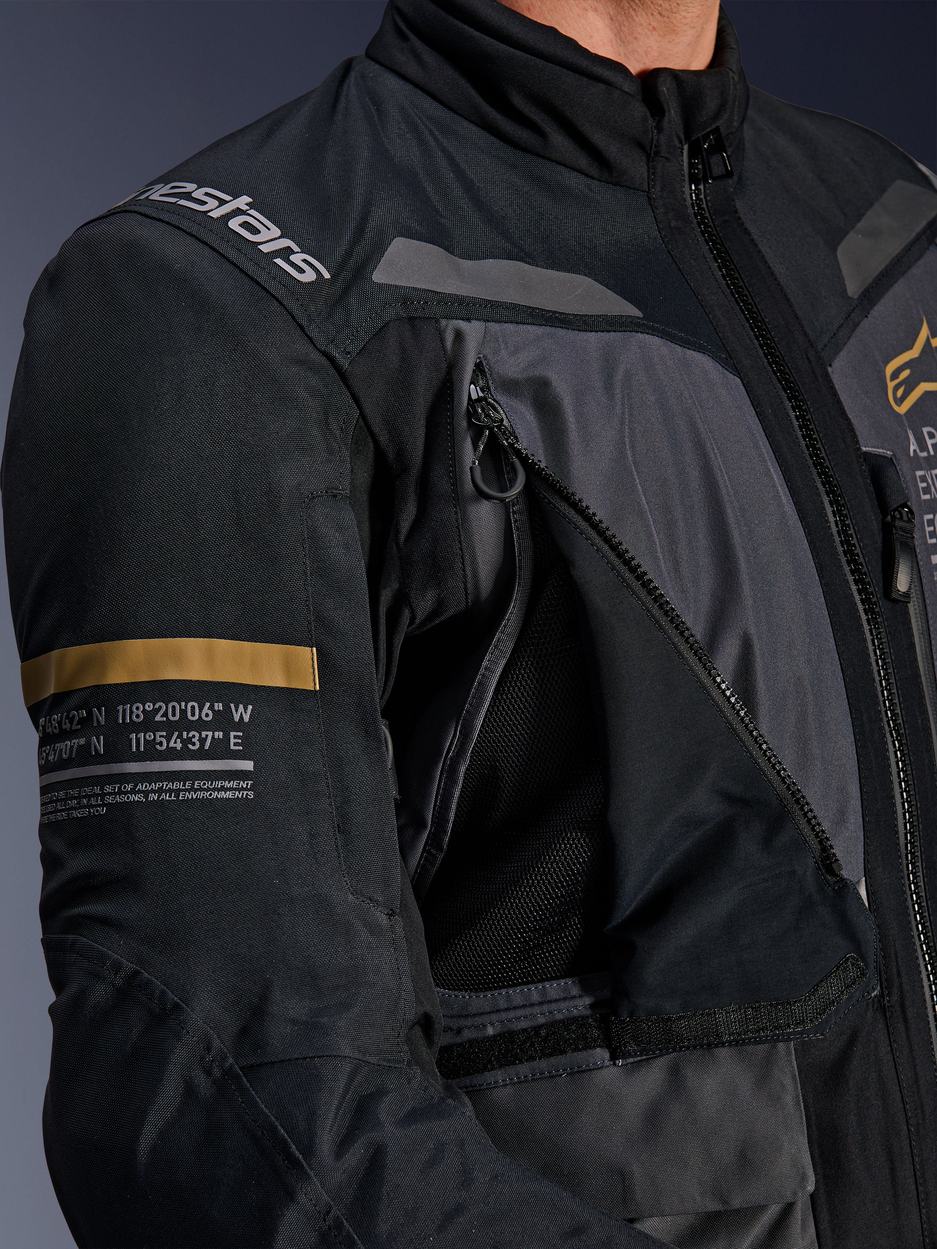 Alpinestars ST-7 2L GORE-TEX Jacke, Dunkelgrau und Schwarz, Nahaufnahme mit geöffnetem Belüftungspanel mit Reißverschluss und Mesh an der Brust, Alpinestars-Logo auf der Schulter und khakifarbenem Streifen auf dem Ärmel mit aufgedruckten geografischen Koordinaten, technische Adventure-Touring-Motorradausrüstung