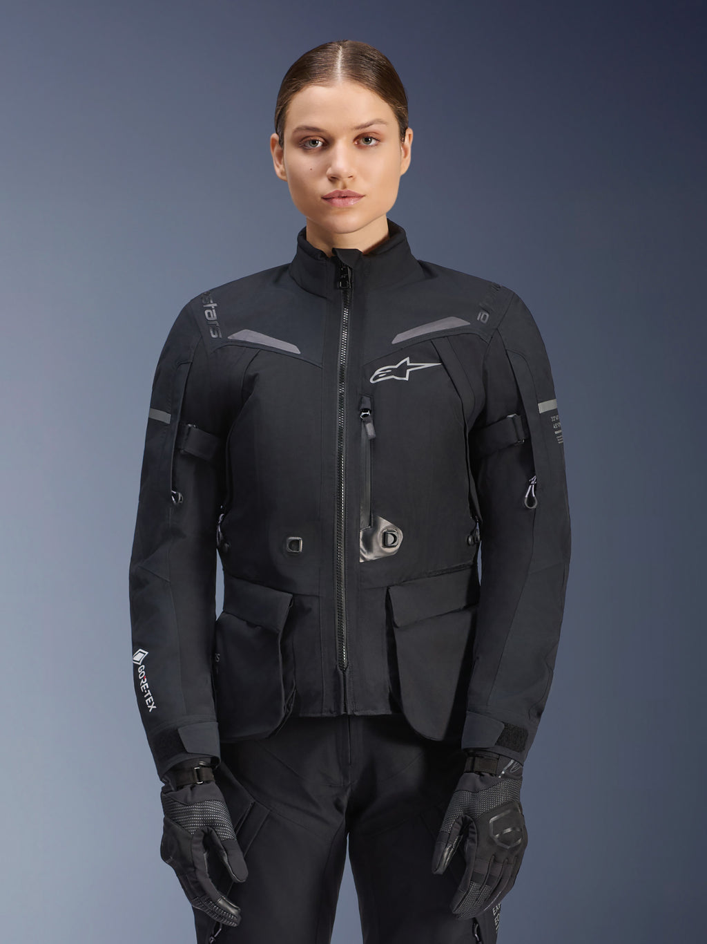 Frau in Alpinestars Stella ST-7 2L GORE-TEX Jacke, Schwarz mit dunkelgrauen Akzenten, Motorrad-Tourenjacke mit großen Cargo-Taschen und hohem Kragen, kombiniert mit schwarzen Handschuhen und Hosen, wasserdichte Adventure-Fahrbekleidung