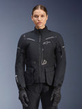 Frau in Alpinestars Stella ST-7 2L GORE-TEX Jacke, Schwarz mit dunkelgrauen Akzenten, Motorrad-Tourenjacke mit großen Cargo-Taschen und hohem Kragen, kombiniert mit schwarzen Handschuhen und Hosen, wasserdichte Adventure-Fahrbekleidung