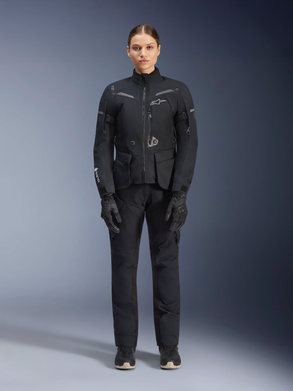 Motorradfahrerin in Alpinestars Damen Stella ST-7 2L GORE-TEX Jacke und Hose, schwarzes Motorrad-Tourenoutfit, wasserdichte und atmungsaktive technische Ausrüstung, mit passenden schwarzen Handschuhen und Stiefeln, Ganzkörper-Schutzausrüstung mit reflektierenden Elementen, modernes technisches Design für Adventure-Riding