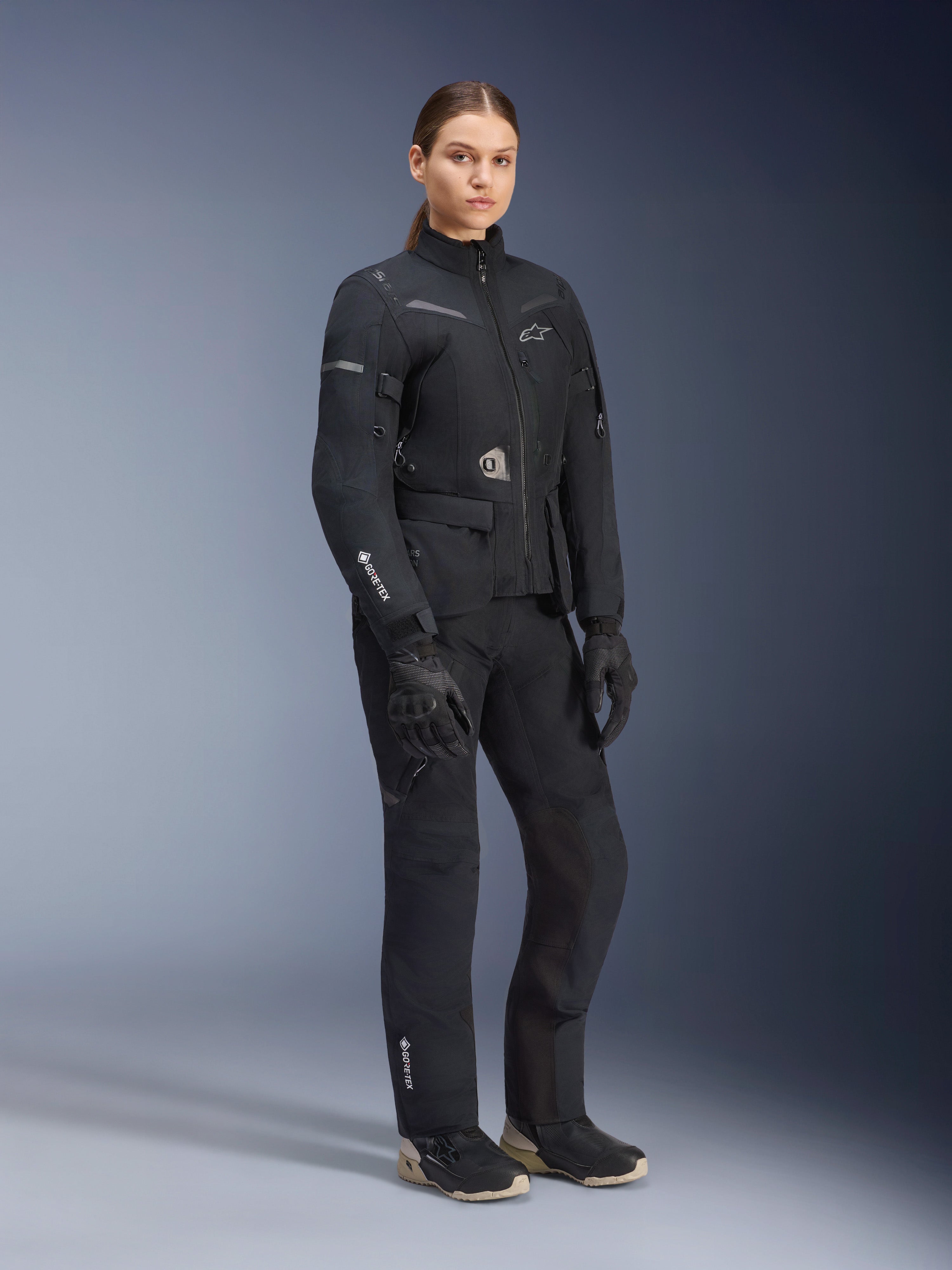 Weibliches Modell im Alpinestars Stella ST-7 2L GORE-TEX Adventure-Tourenanzug, Jacke und Hose in Schwarz und Dunkelgrau, mit markantem GORE-TEX-Branding auf Ärmel und Bein, kombiniert mit schwarzen Handschuhen und Motorradstiefeln, professionelle Motorradausrüstung für Touren bei jedem Wetter.
