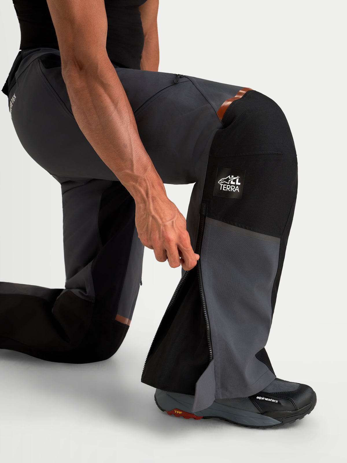 Alpinestars St 7 V 2 2 L Gore Tex Hose, Herren-Adventure-Motorradhose in Dunkelgrau und Schwarz, Nahaufnahme eines knienden Modells mit geöffnetem Unterschenkel-Reißverschluss und All Terra-Branding