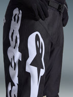 Alpinestars 2026 Racer Air Riway Pants, black motocross pants with bold white Alpinestars logo on the thigh, close-up showing technical fabric and adjustable waist strap, worn with black gloves for off-road riding