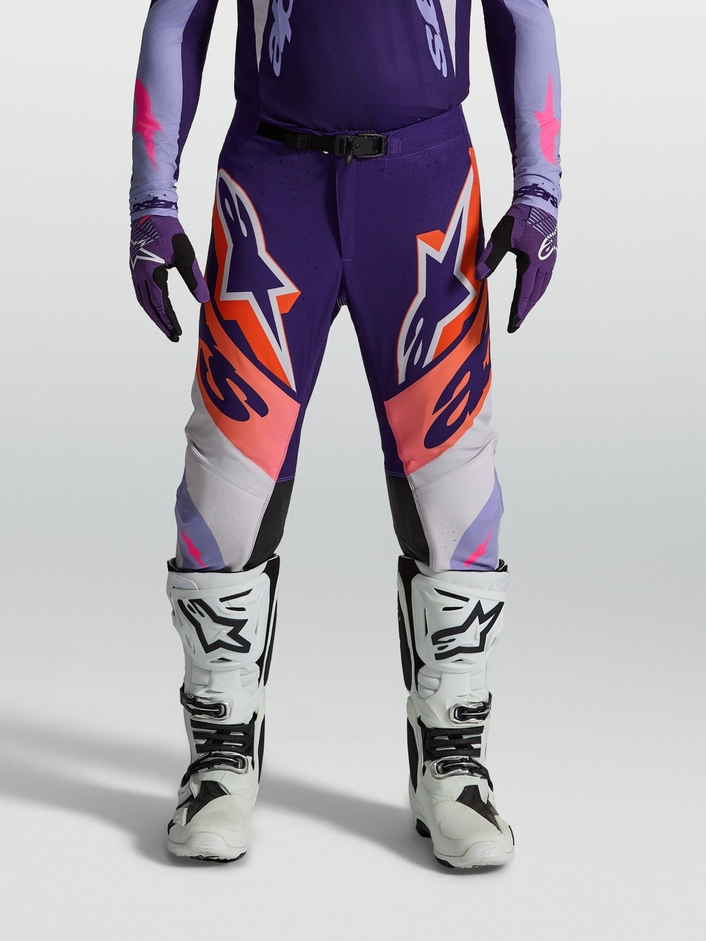 Alpinestars Supertech Dyna Hose, Motocrosshose für Herren, Lila und Pink Fluo mit orangen Details, athletische Rennpassform aus Stretch-Ripstop, getragen mit weißen Stiefeln für den Offroad-Einsatz.