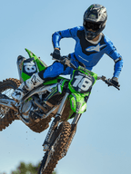Motocross-Fahrer mitten im Sprung auf einem grünen Motorrad, trägt Alpinestars 2025 Racer Veil Hose in Blau und Schwarz, passendes blaues Trikot mit weißem Brustlogo, weiß-schwarze Stiefel und schwarzen Helm, Hochleistungs-Offroad-Ausrüstung für den Rennsport.