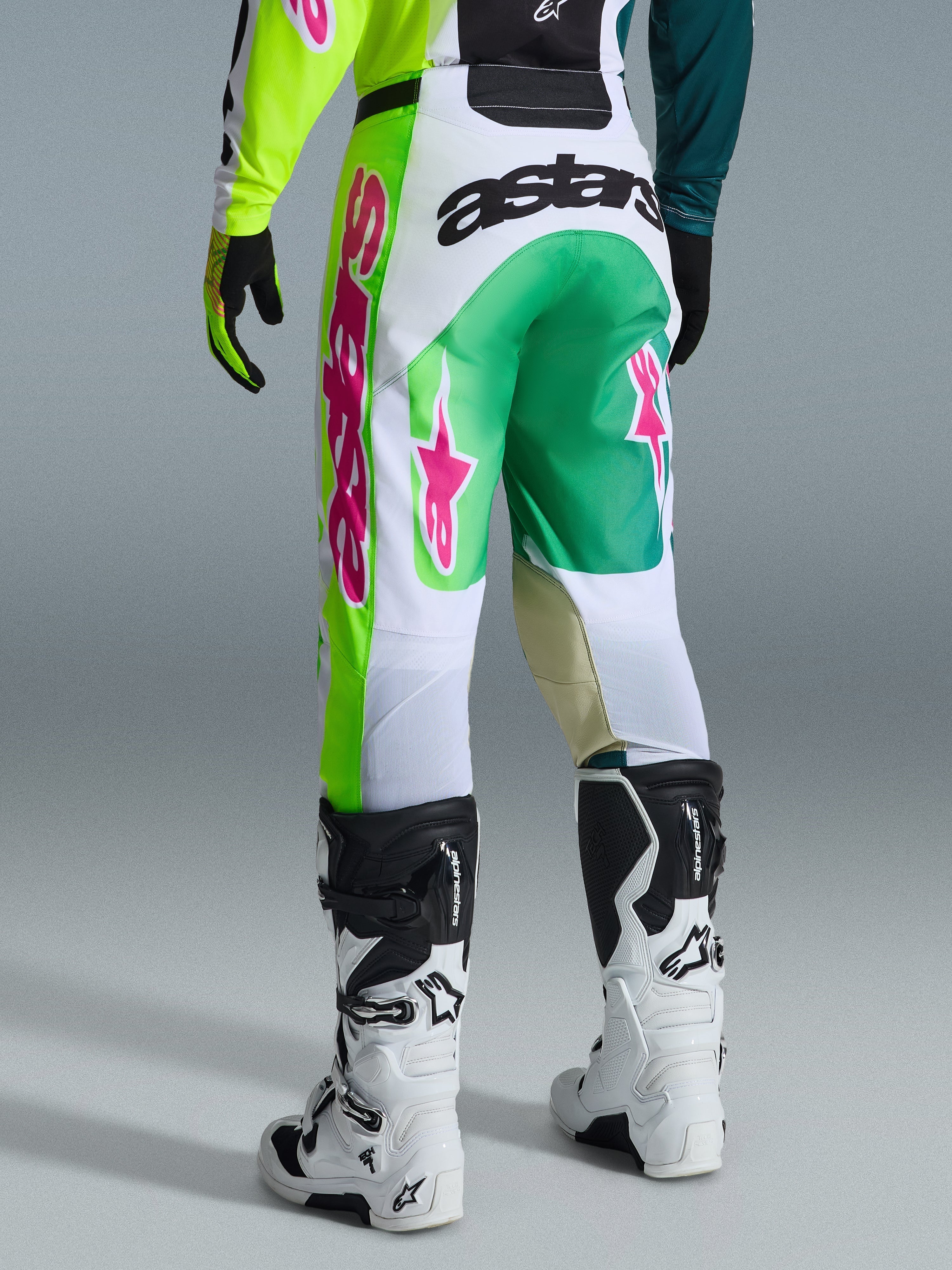 Alpinestars 2026 Racer Portl Hose, Motocross-Hose, Grün, Weiß und Schwarz mit pinken Akzenten, Rückansicht mit markantem „astars“-Logo am Gesäß und pinken Stern-Logos auf den Beinen, kombiniert mit weiß-schwarzen Motocross-Stiefeln, Performance-Ausrüstung für den Offroad-Sport