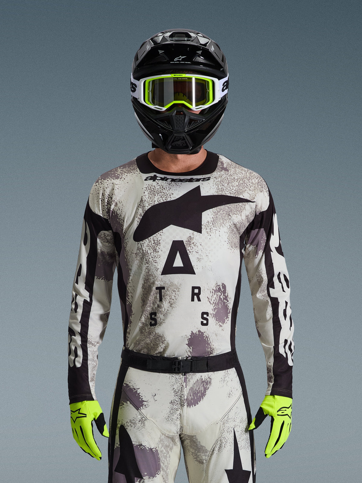 Motocross-Fahrer im Alpinestars 2026 Racer Lahnd Jersey, Sand-Camo-Muster in Grau und Weiß, großes schwarzes Astar-Logo und Branding auf der Brust, kombiniert mit einem schwarzen Helm, neongelber Brille und Handschuhen, Performance-Ausrüstung für Offroad-Fahrten