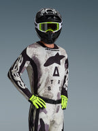 Motocross-Fahrer im Alpinestars 2026 Racer Lahnd Trikot und Bekleidungsset in Sand Camo, hellgraues Camouflage-Muster mit schwarzem Logo-Branding, schwarzer Helm mit weiß-neongelber Brille, leuchtend neongelbe Handschuhe, professionelle Offroad-Rennbekleidung