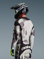 Motocross-Fahrer von hinten im Alpinestars 2026 Racer Lahnd Bekleidungsset, Trikot und Hose in Grau und Weiß Sand Camo mit schwarzen Akzenten, schwarzer Helm mit weißem Alpinestars-Logo, neongelbe und schwarze Handschuhe, abgestimmtes Offroad-Performance-Outfit