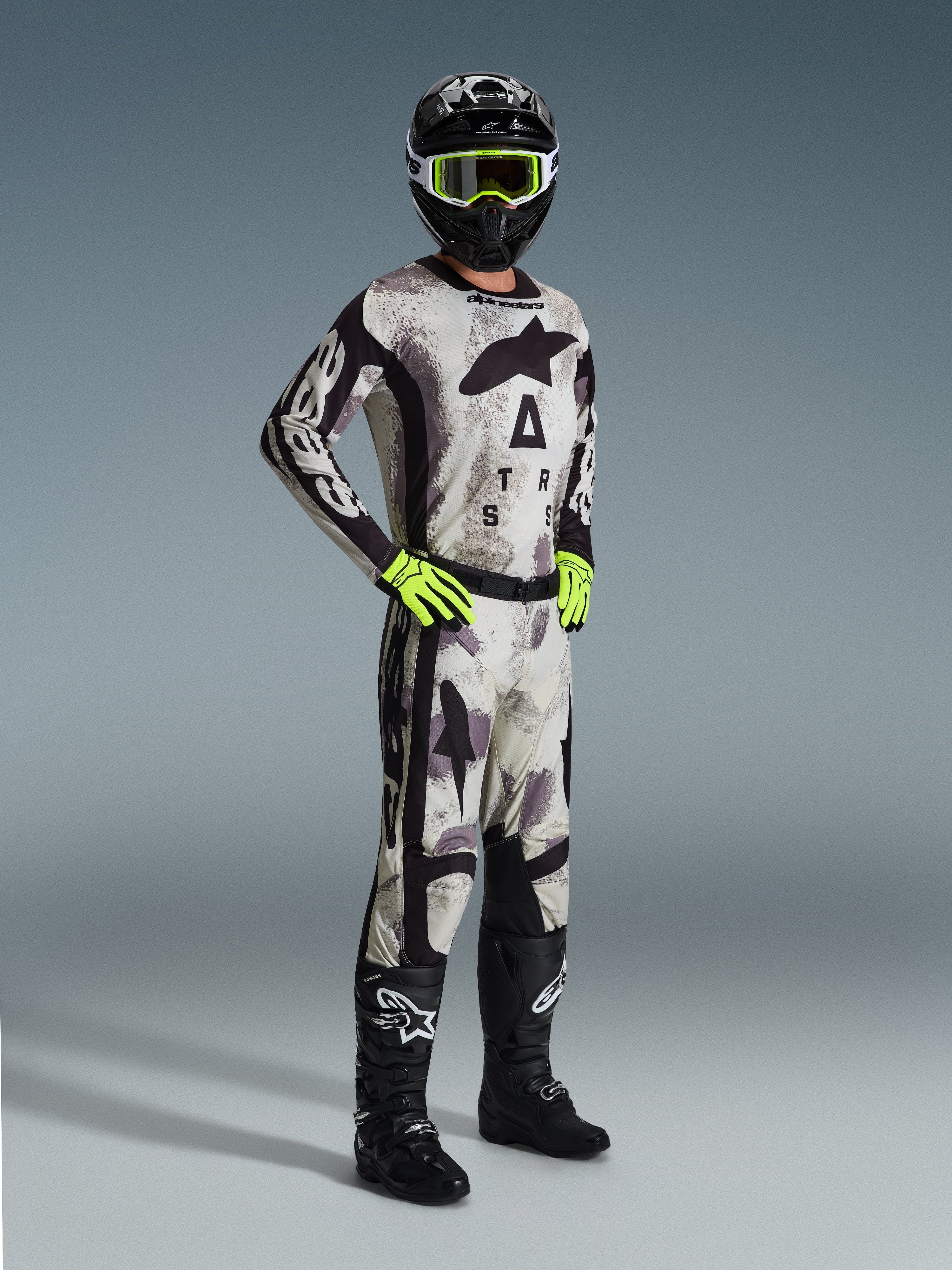 Motocross-Fahrer im 2026 Racer Lahnd Trikot in Sand Camo, grau-schwarzes Camouflage-Muster mit Alpinestars-Branding, passende Motocross-Hose, neongelbe Handschuhe, schwarzer Helm mit gelber Brille und schwarze Stiefel, komplette professionelle Offroad-Rennausrüstung.