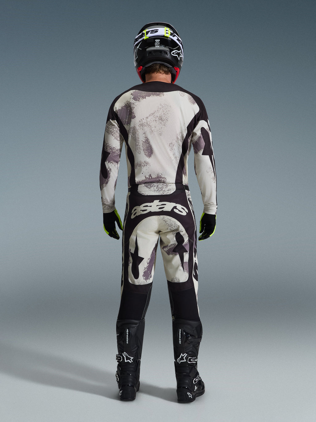 Motocross-Fahrer in komplettem Alpinestars 2026 Racer Lahnd Ausrüstungsset, von hinten gesehen, Jersey und Hose in Sand Camo mit hellgrauem und schwarzem abstraktem Muster, schwarzer Helm, schwarz-neongelbe Handschuhe, schwarze Stiefel, mit 'astars'-Branding auf der Rückseite der Hose