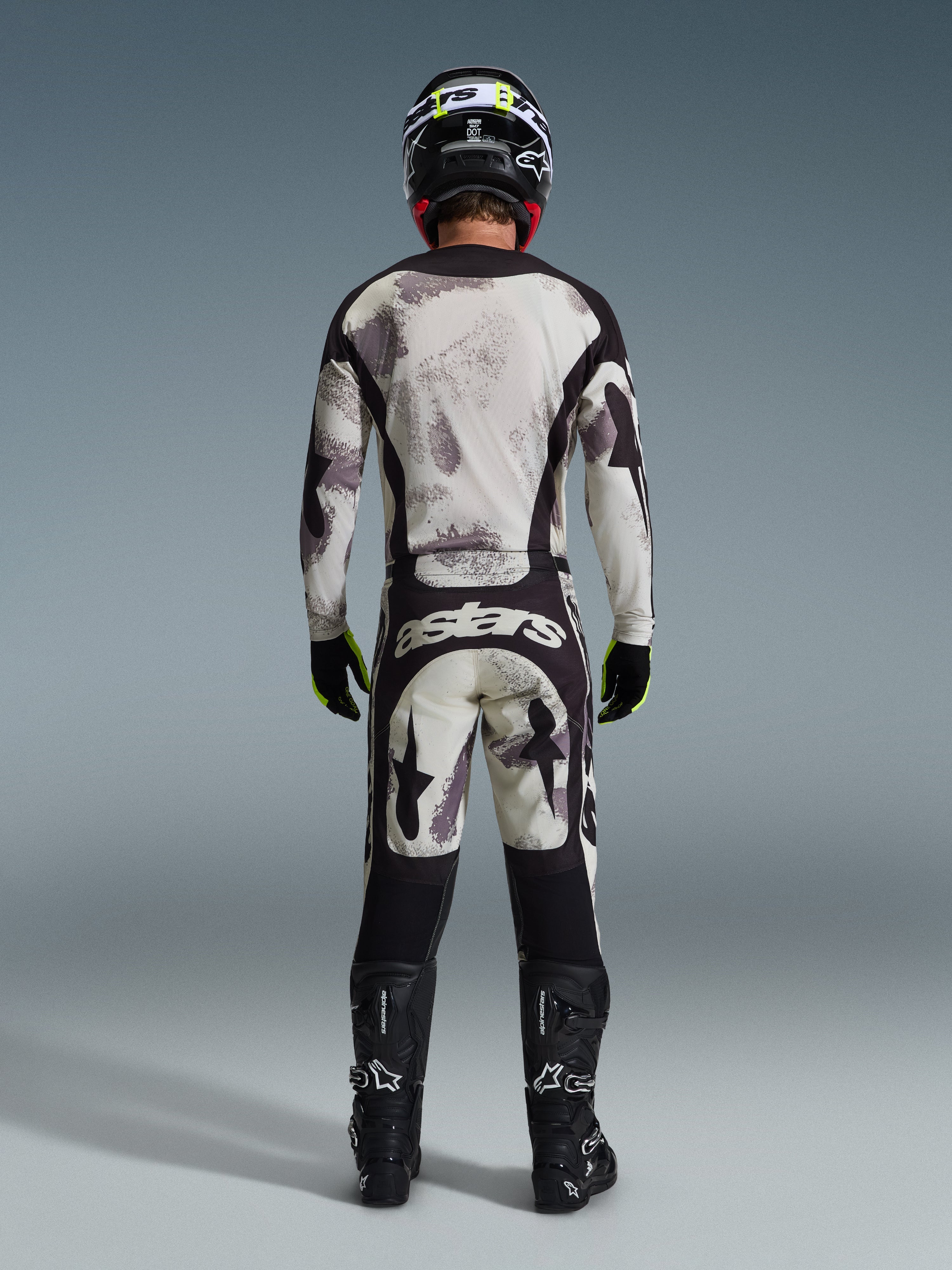 Motocross-Fahrer in komplettem Alpinestars 2026 Racer Lahnd Ausrüstungsset, von hinten gesehen, Jersey und Hose in Sand Camo mit hellgrauem und schwarzem abstraktem Muster, schwarzer Helm, schwarz-neongelbe Handschuhe, schwarze Stiefel, mit 'astars'-Branding auf der Rückseite der Hose
