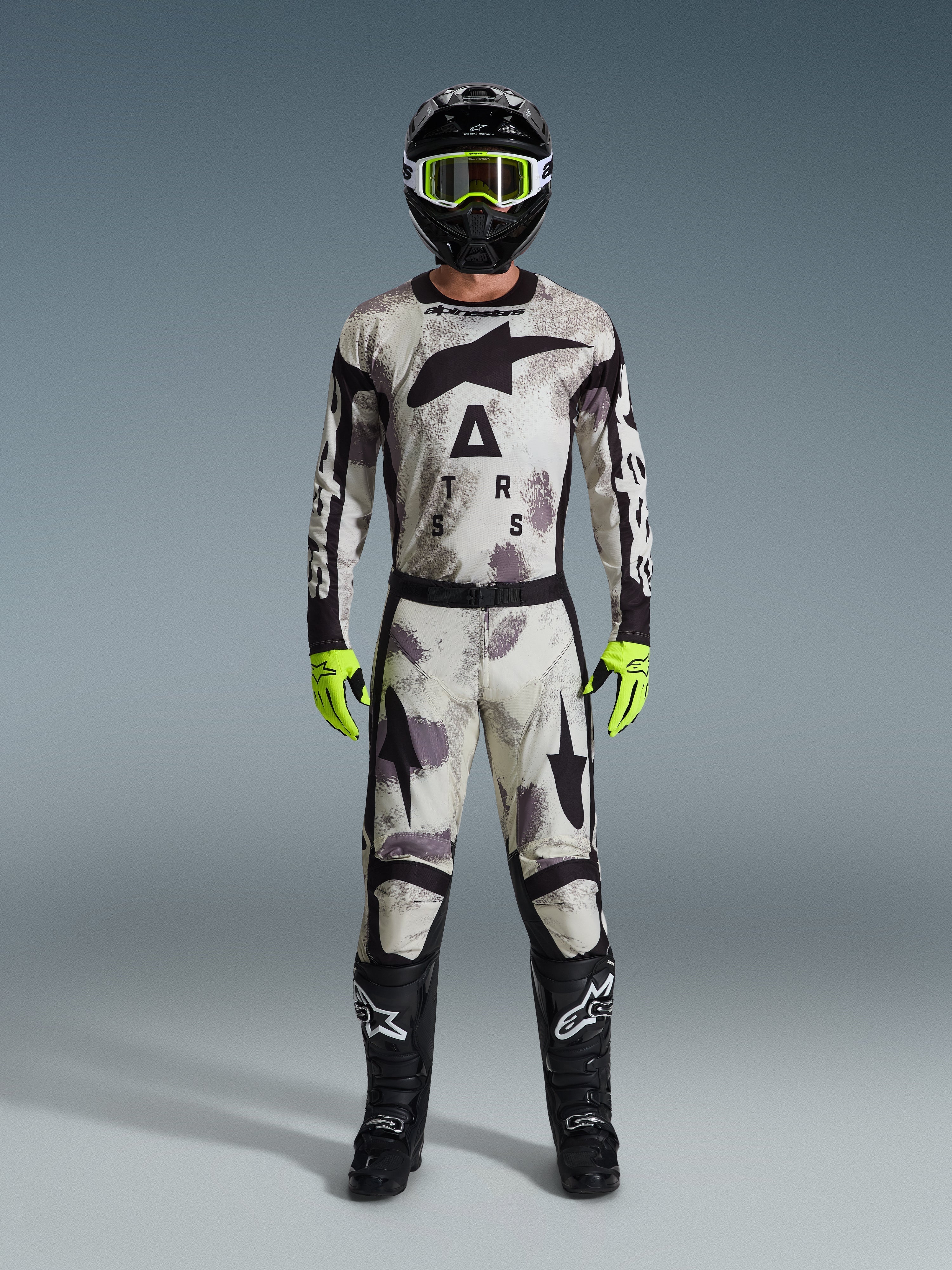 Motocross-Fahrer im Alpinestars 2026 Racer Lahnd Gear Set, Anzug mit Sand-Camo-Muster in Weiß, Grau und Schwarz, schwarzer Helm mit neongrüner Brille, leuchtend neongrüne Handschuhe und schwarze Stiefel mit weißen Logos, komplettes koordiniertes Outfit für den Offroad-Rennsport