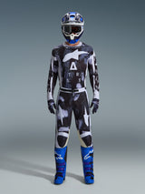 Motocross rider wearing Alpinestars 2026 Racer Lahnd Pants in Iron Camo, gray and black camouflage design, matching jersey, blue and white helmet with goggles, and bright blue boots, coordinated motocross gear set.