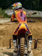 Motocross-Fahrer von hinten in Alpinestars 2025 Supertech Lipan Ausrüstungsset, Gelb und Pink, Trikot mit Nummer 1 und „LAWRENCE“ auf dem Rücken, gelbe Hose mit markantem pinkem „ASTARS“-Branding, weiße Stiefel und passender Helm, sitzend auf einem Motocross-Bike am Startgatter