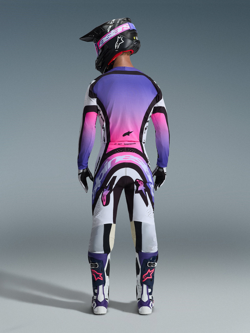 Motocross-Fahrer in der Rückansicht, komplette Rennausrüstung, Trikot mit lila-pinkem Farbverlauf, Alpinestars 2026 Supertech AFD Vista Hose in Weiß, Lila und Schwarz, schwarzer Helm mit lila Brille, weiß-lila Stiefel, farblich abgestimmtes Offroad-Rennset.