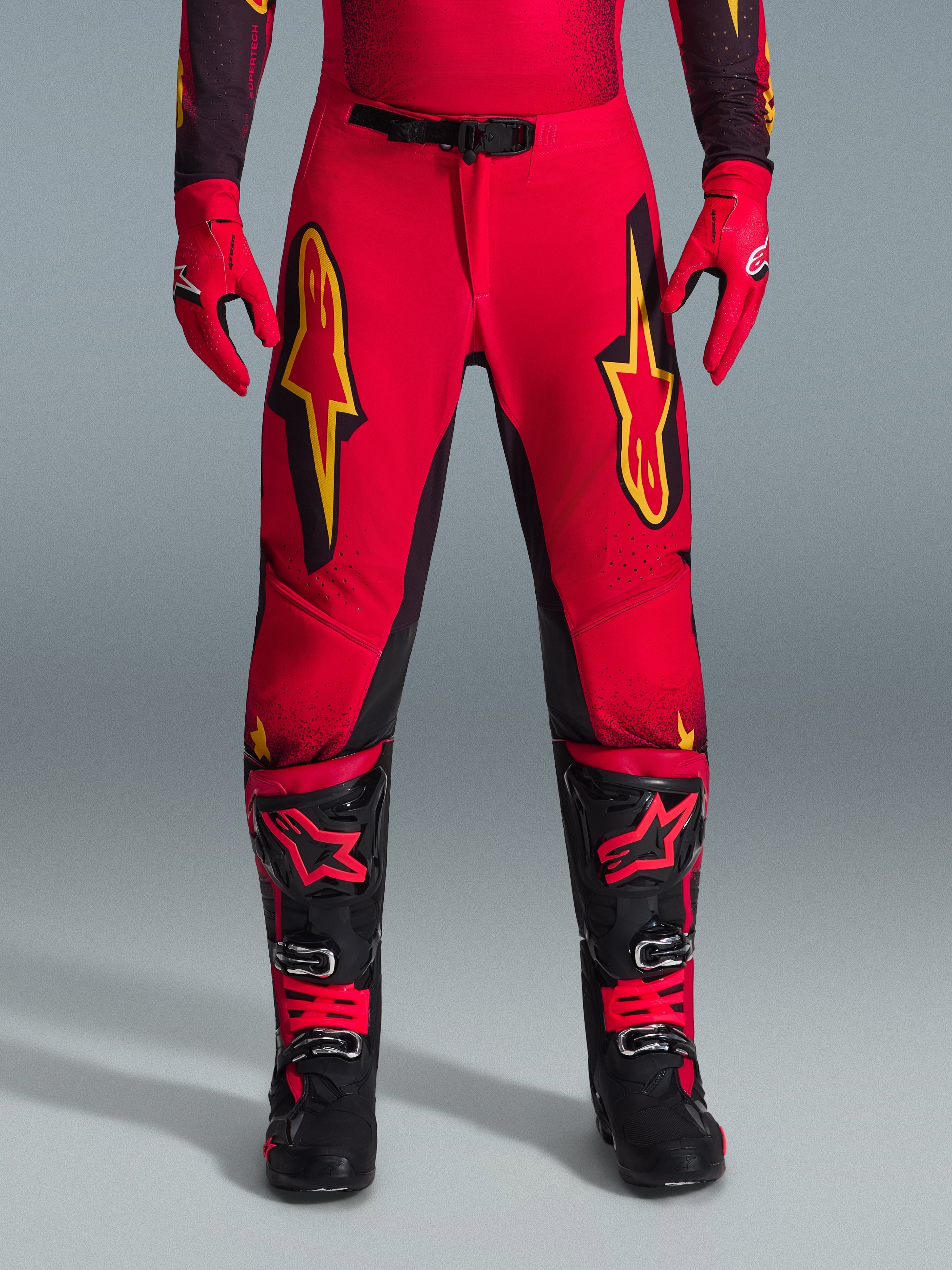 Alpinestars 2026 Supertech Scenz Hose, Motocross-Hose, Rot, Schwarz und Gelb, athletische Profi-Passform mit großen gelben Stern-Logos auf den Oberschenkeln, kombiniert mit passenden roten Handschuhen und schwarz-roten technischen Stiefeln, entwickelt für Elite-Offroad-Performance