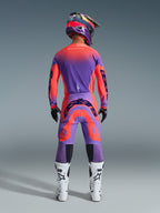 Alpinestars 2026 Supertech Scenz Hose, Motocross-Hose, Hot Coral und Lila, Rückansicht eines Motocross-Fahrers in kompletter Rennausrüstung, mit lebhaftem Farbverlauf-Design und markanten Logos auf der Rückseite, kombiniert mit passendem Jersey, Helm und weißen Stiefeln.