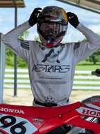 Motocross-Fahrer in Alpinestars 2025 Racer Hollow Ausrüstung, helles Camo-Trikot mit markantem ASTARS-Branding, justiert einen Red Bull Helm mit klarer Brille, neben einem roten Honda-Dirtbike in einer Offroad-Umgebung.
