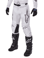 Alpinestars 2025 Racer Hollow Hose, Motocross-Hose, Weiß und Grau Camouflage, mit großen schwarz umrandeten Stern-Logos und schwarzen Leder-Innenknie-Einsätzen, kombiniert mit schwarzen Motocross-Stiefeln und Handschuhen, strapazierfähiges Design für Offroad-Rennsport