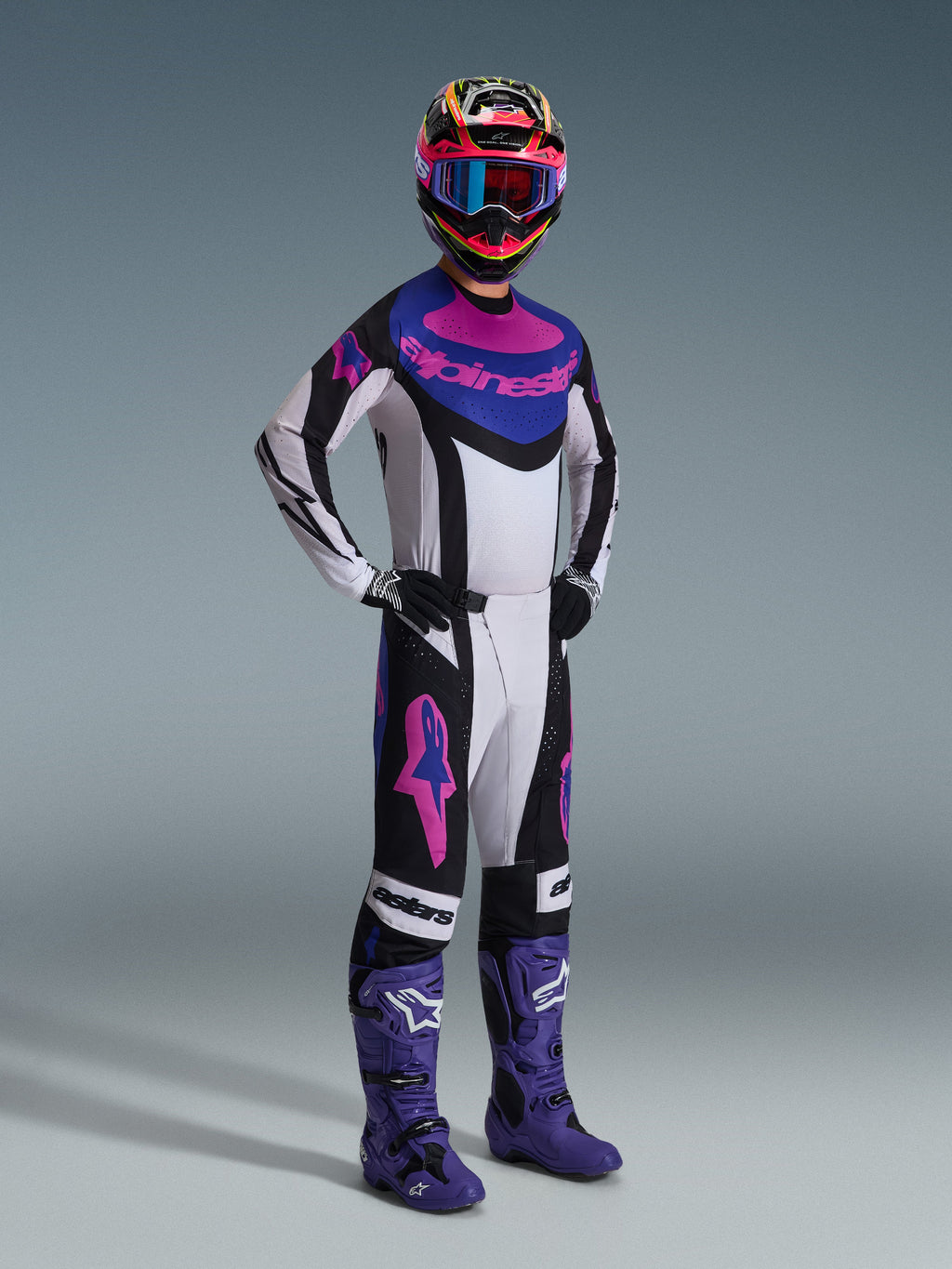 Motocross-Fahrer im Alpinestars 2026 Techstar Knif Gear Set, Schwarz, Hellgrau und Lila, weißes Trikot mit blauen und rosa Akzenten, passende Hose mit lila Stern-Logos, farbenfroher pinker und gelber Helm, lila Motocross-Stiefel und schwarze Handschuhe, professionelle Ausrüstung für den Offroad-Rennsport.