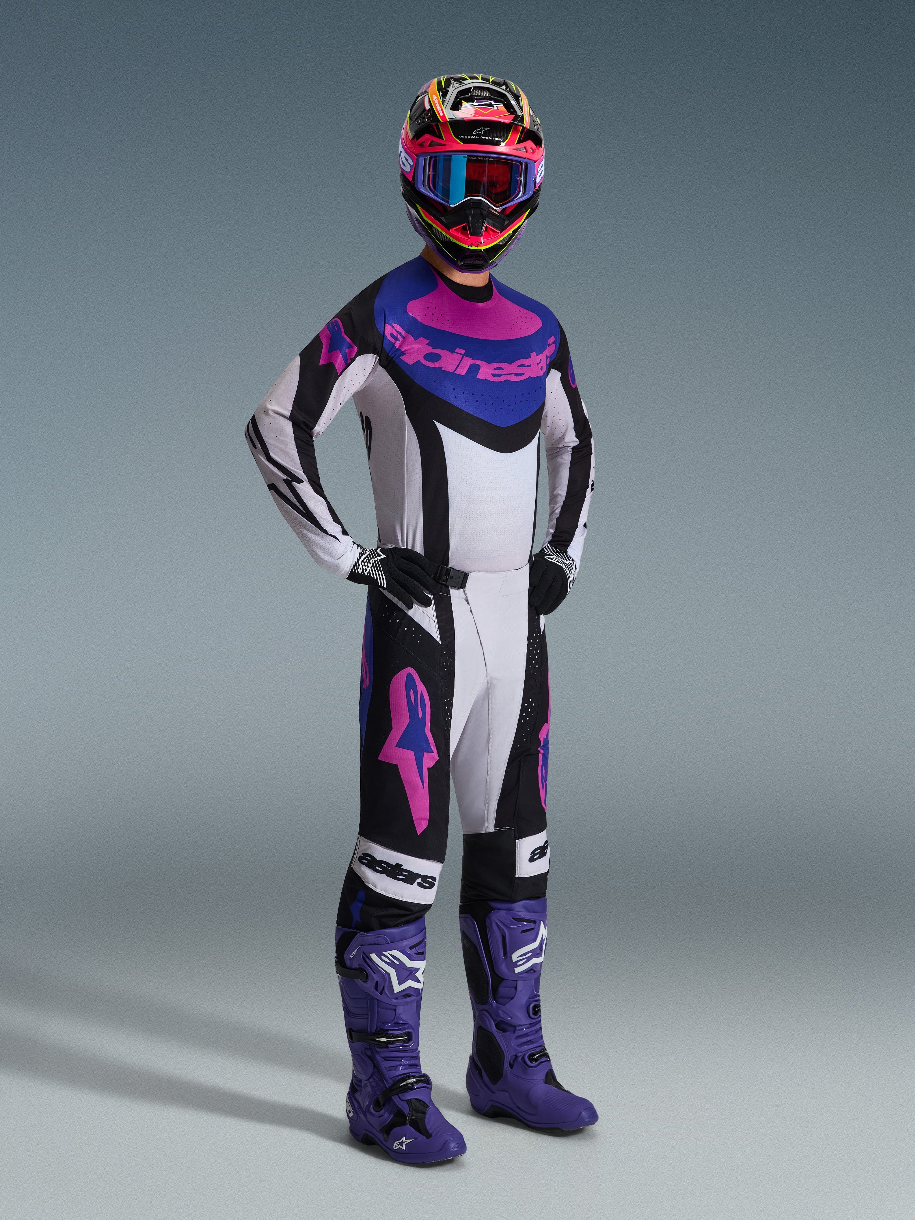 Motocross-Fahrer im Alpinestars 2026 Techstar Knif Gear Set, Schwarz, Hellgrau und Lila, weißes Trikot mit blauen und rosa Akzenten, passende Hose mit lila Stern-Logos, farbenfroher pinker und gelber Helm, lila Motocross-Stiefel und schwarze Handschuhe, professionelle Ausrüstung für den Offroad-Rennsport.