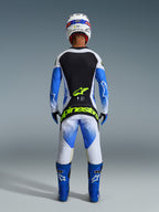 Motocross-Fahrer von hinten in kompletter Rennausrüstung, 2026 Techstar Knif Hose in UCLA Blue Black und Yellow Fluo, passendes Langarm-Trikot mit schwarz-weißen Einsätzen, blaue Offroad-Stiefel, weißer Helm, mit markantem neongelbem Alpinestars-Branding am Gesäß.