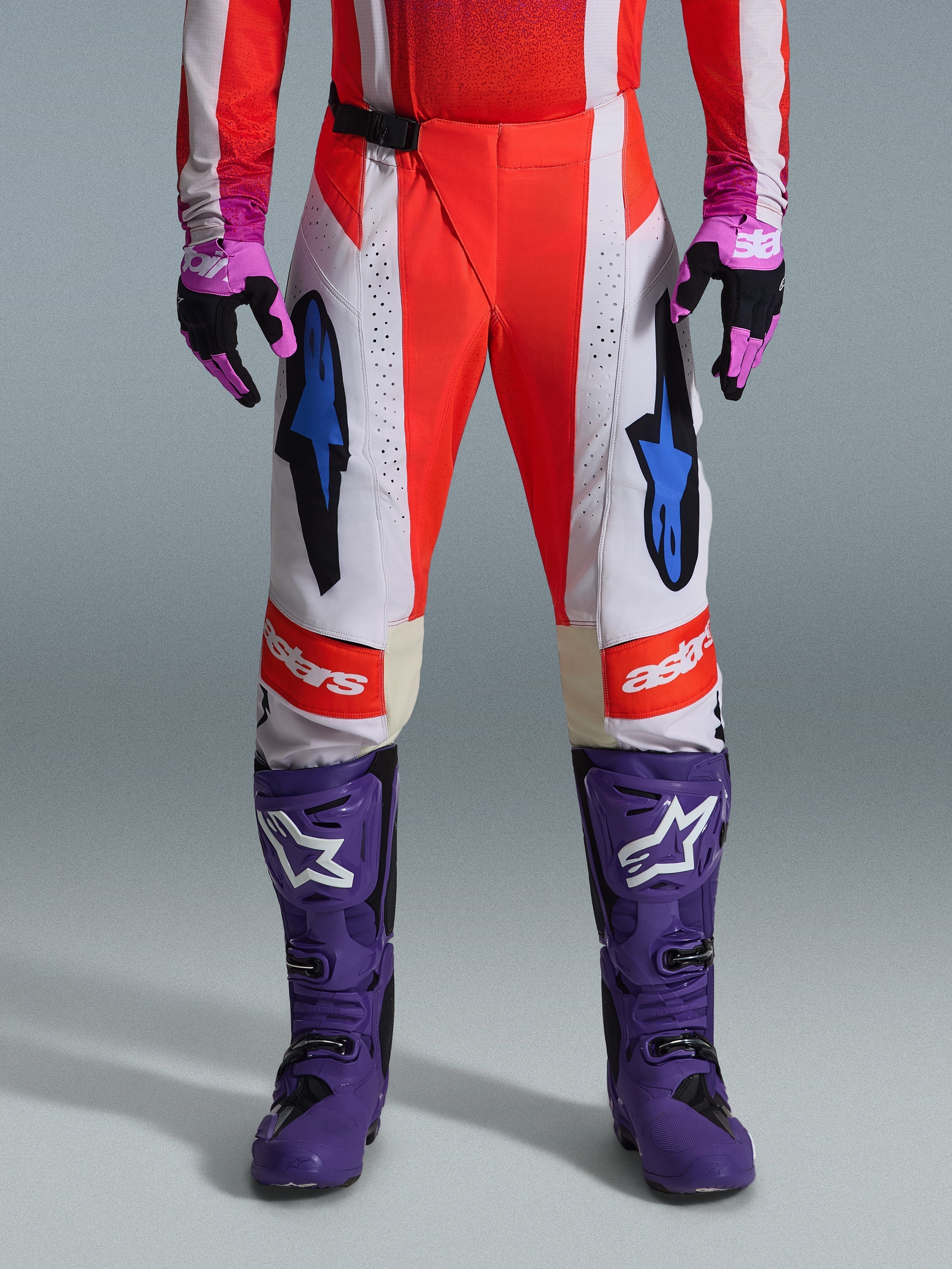 Alpinestars 2026 Techstar Knif Hose, Motocross-Hose in Orange Fluo und Hellgrau mit schwarzen Akzenten, kombiniert mit lila Motocross-Stiefeln und Handschuhen, mit blauen Stern-Logos und Multi-Panel-Konstruktion für Offroad-Performance