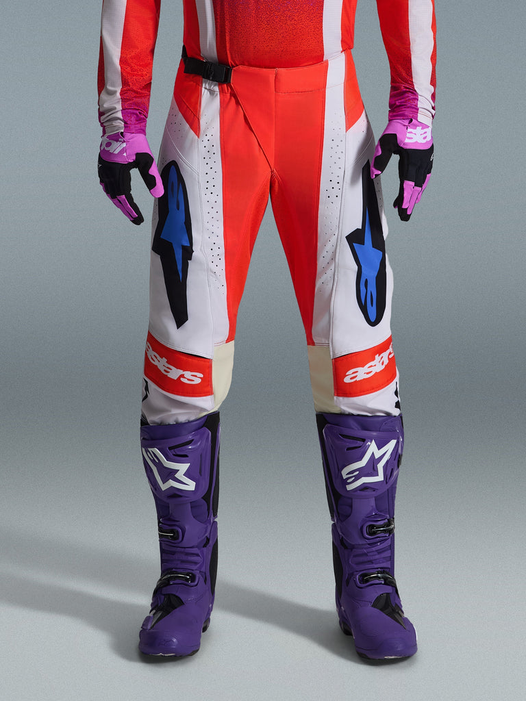 Alpinestars 2026 Techstar Knif Hose, Motocross-Hose in Orange Fluo und Hellgrau mit schwarzen Akzenten, kombiniert mit lila Motocross-Stiefeln und Handschuhen, mit blauen Stern-Logos und Multi-Panel-Konstruktion für Offroad-Performance