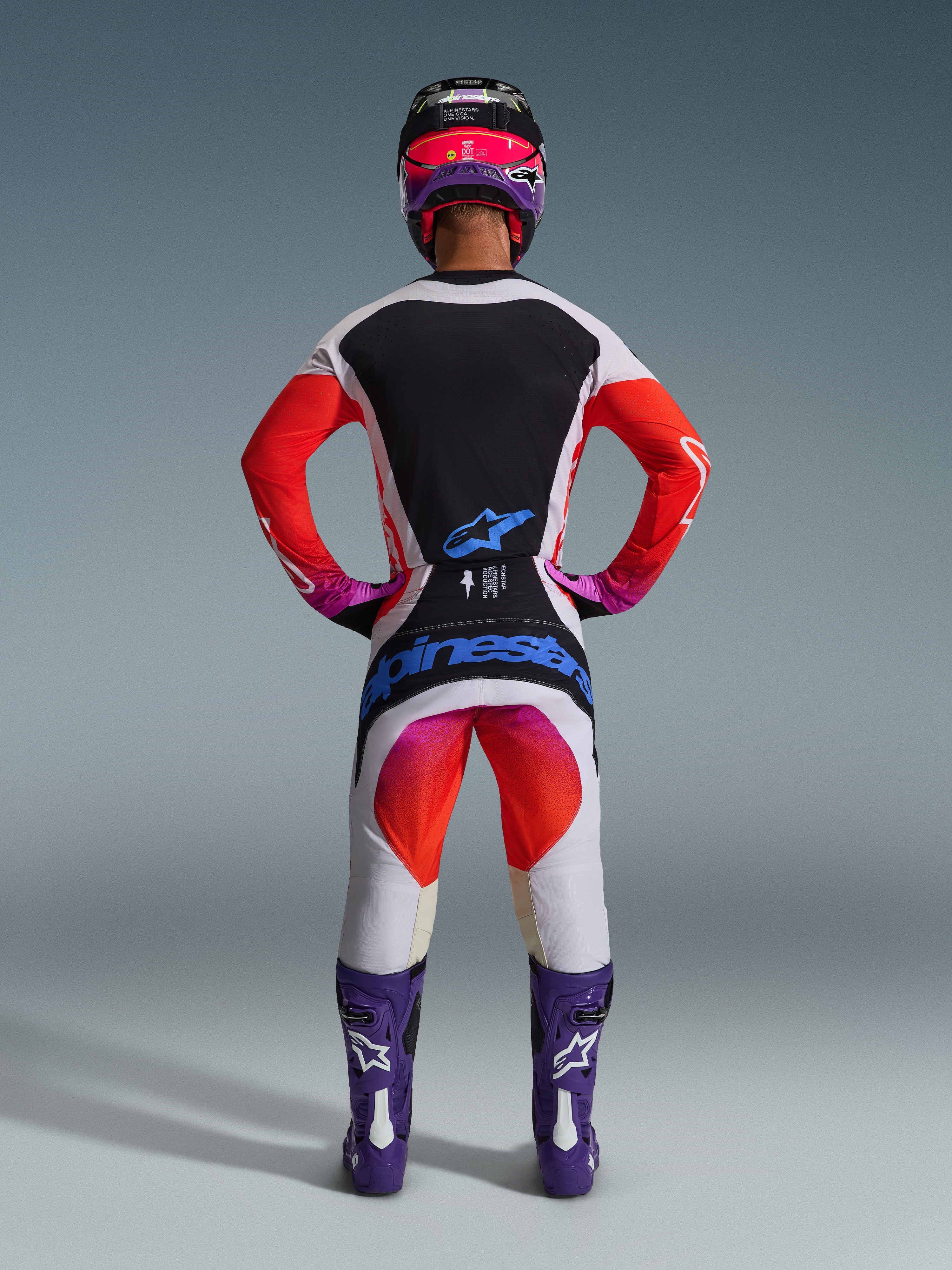 Motocross-Fahrer in kompletter 2026 Techstar Knif Rennausrüstung von hinten, hellgraues Trikot mit schwarzem Rückeneinsatz und orangefarbenen Ärmeln, passende weiß-orangefarbene Hose mit blauem Alpinestars-Branding, lila Stiefel und schwarz-pinker Helm, abgestimmtes professionelles Outfit für den Offroad-Rennsport