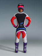 Motocross-Fahrer in der Rückansicht mit 2026 Techstar Knif Hose und passendem Trikot, hellgraues und schwarzes Design mit leuchtend orangefarbenen und lila Akzenten, blaues Alpinestars-Logo auf der Gesäßpartie, lila Stiefel und Helm, professionelles Offroad-Rennausrüstungsset.