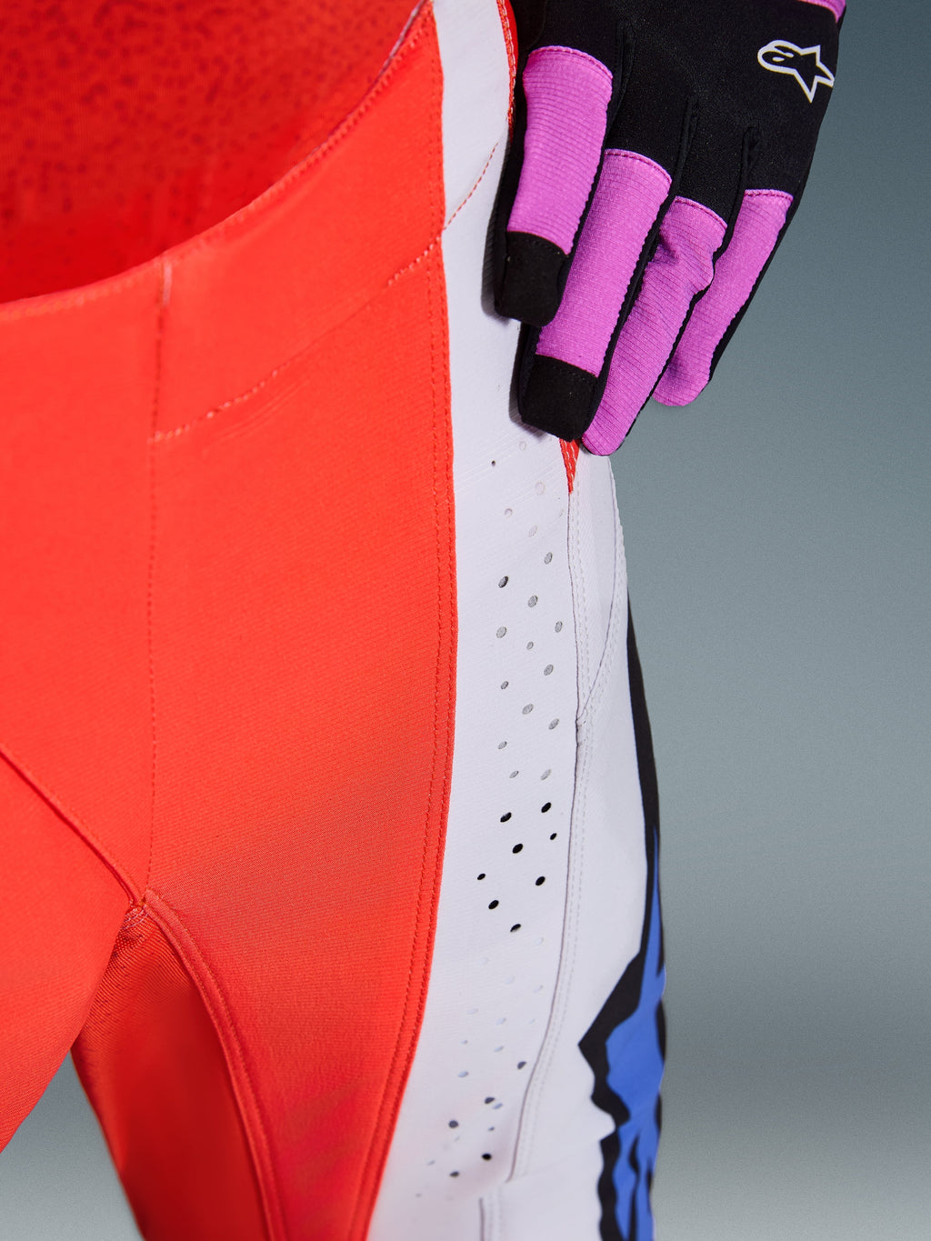 Alpinestars 2026 Techstar Knif Hose, Motocross-Hose, Orange Fluo und Hellgrau mit blauen Akzenten, Nahaufnahme der lasergeschnittenen Belüftungsperforationen am Oberschenkel, kombiniert mit einem schwarz-lila Motocross-Handschuh