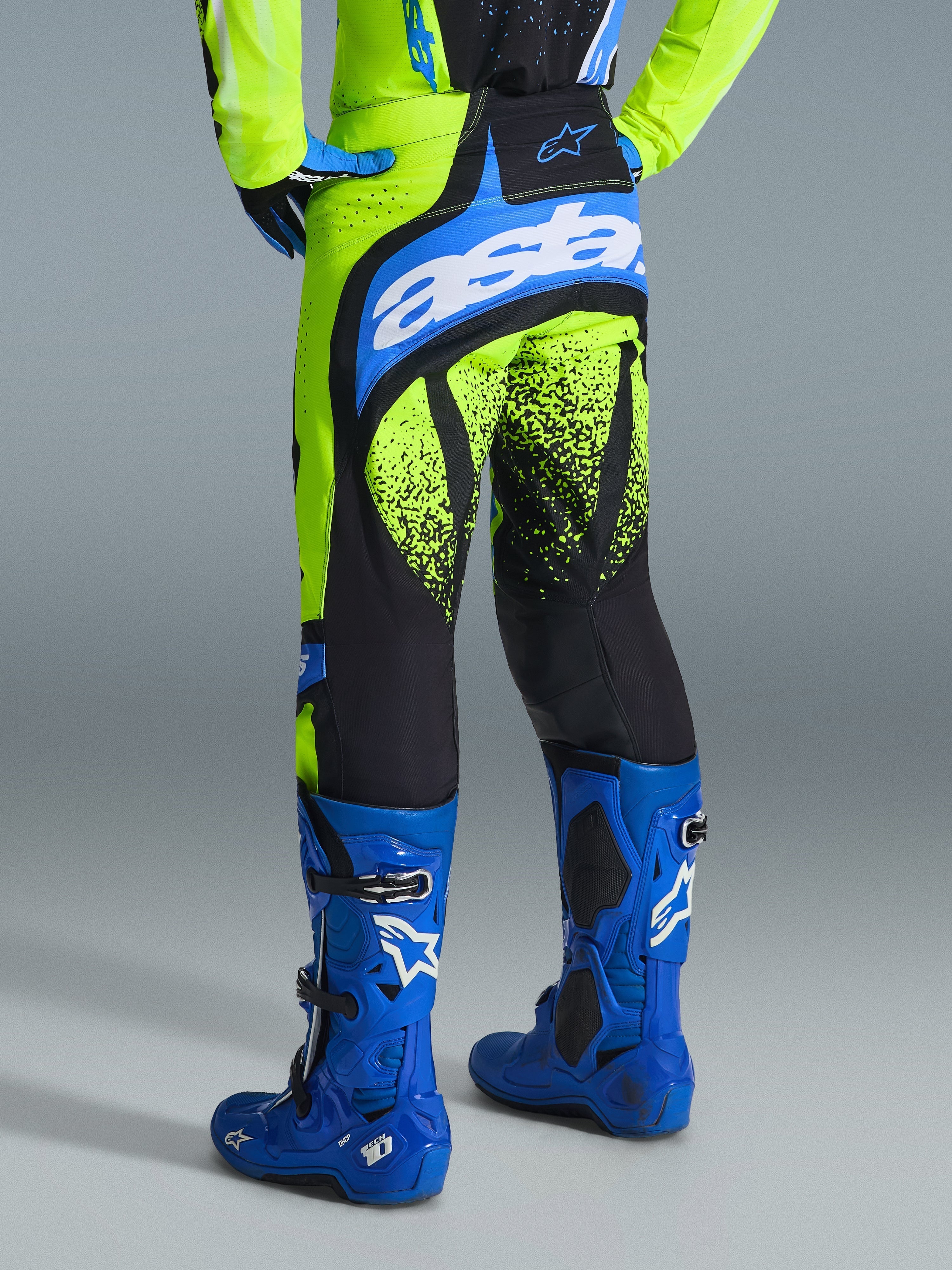 Alpinestars 2026 Techstar Nomur Hose, Motocross-Hose, Gelb Fluo und UCLA Blau, Rückansicht eines Fahrers mit blauen Stiefeln, mit „astars“-Branding auf der Sitzfläche und schwarzen Splatter-Grafiken an den Beinen