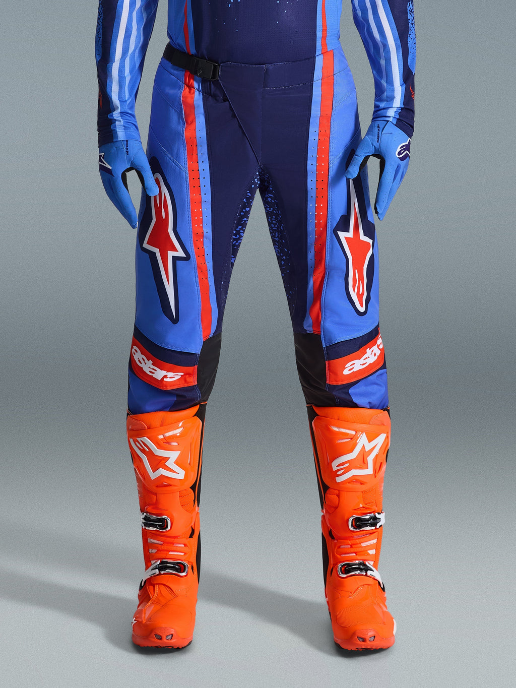 Alpinestars 2026 Techstar Nomur Hose, Motocross-Hose, Dunkelblau, Orange und UCLA-Blau, mit markanten orangefarbenen Streifen und großen weißen Stern-Logos auf den Oberschenkeln, getragen mit leuchtend orangefarbenen Rennstiefeln und blauen Handschuhen, athletische Rennpassform für Offroad-Performance