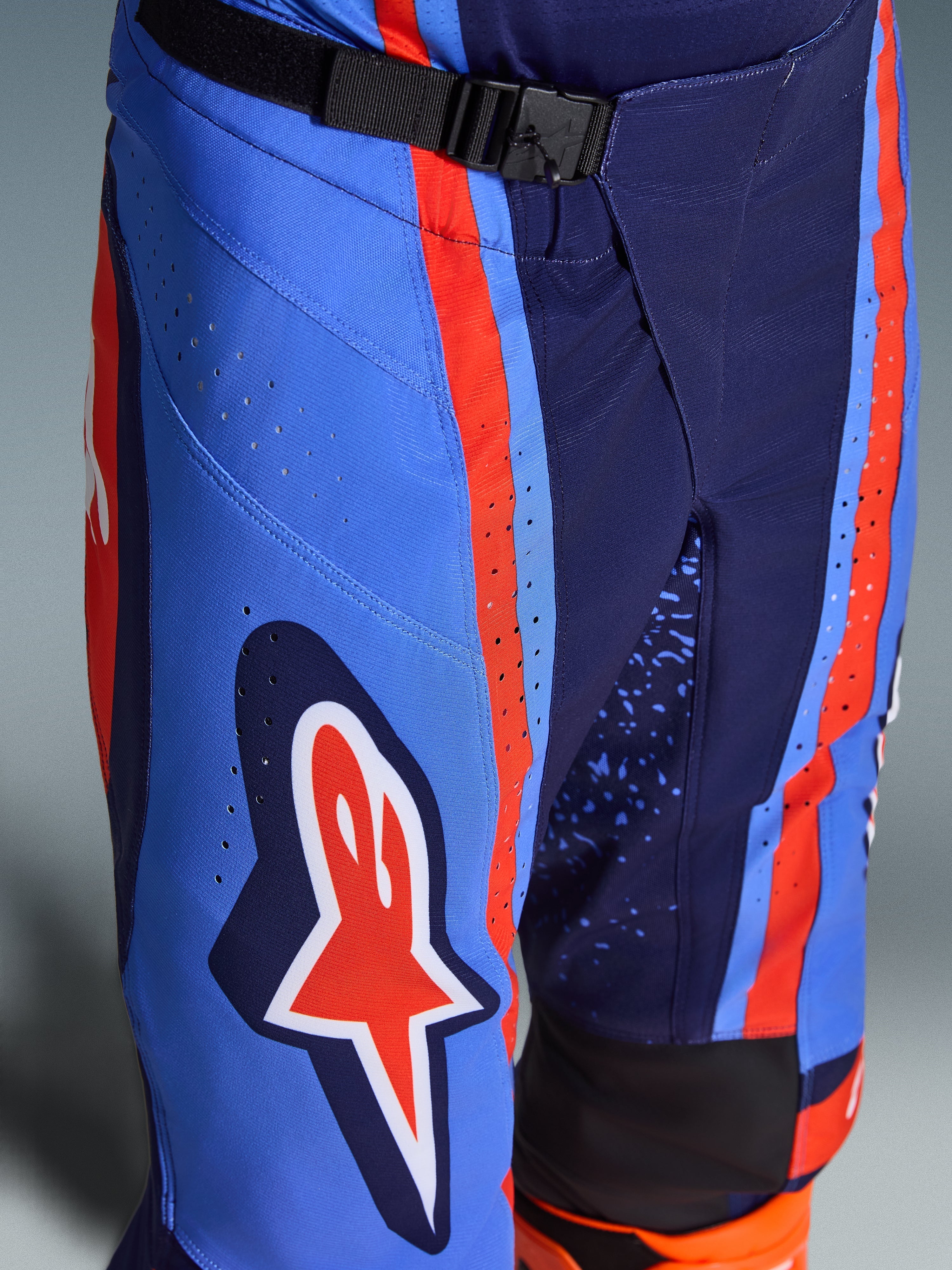 Alpinestars 2026 Techstar Nomur Hose, Motocross-Hose in Dark Navy Orange und UCLA Blue, Nahaufnahme des SX2-Magnetverschlusses am Bund, lasergeschnittene Perforationen am Oberschenkel, ikonisches Stern-Logo, strapazierfähige Offroad-Rennausrüstung.